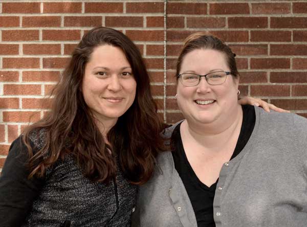 Two women standing side by side in front of a brick wall.