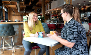 Two women sitting at a table at a coffee shop holding coffee mugs.