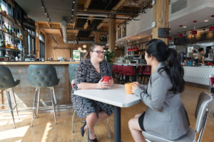 Two women sitting at a table at a coffee shop holding coffee mugs.