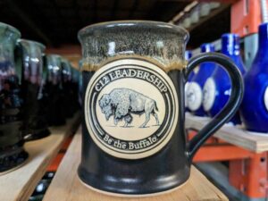 Black handmade coffee mug with clay medallion with illustration of a bison with the text "C12 Leadership. Be the buffalo."
