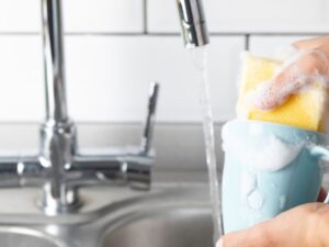person cleaning a blue coffee mug under running water with soap and a sponge