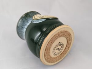green coffee mug laying on its side with the bottom of the mug shown with a cork pad attached, label on the bottom of the mug reads 'handmade grey fox pottery'