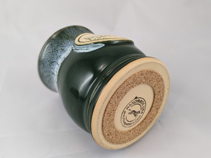 green coffee mug laying on its side with the bottom of the mug shown with a cork pad attached, label on the bottom of the mug reads 'handmade USA greyfoxpottery.mom'